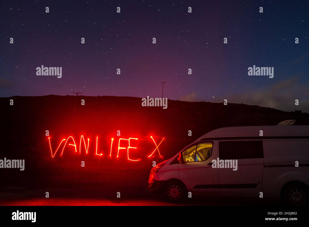 A long exposure light painting of a campervan parked at night with the word vanlife written in red light. Starry night sky Stock Photo
