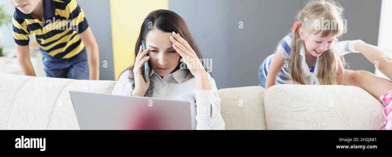 Sad young woman sitting on sofa with laptop and holding her head near children playing at home Stock Photo