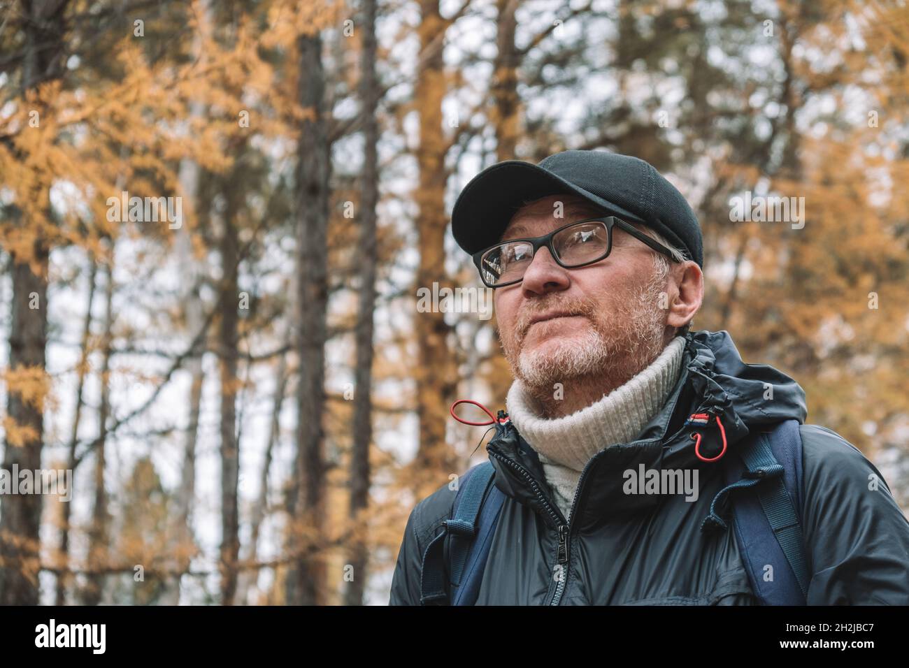 A candid portrait of serious unshaven middle-aged man in glasses and a cap against the background of an autumn forest. Local travel and weekend walks. Stock Photo