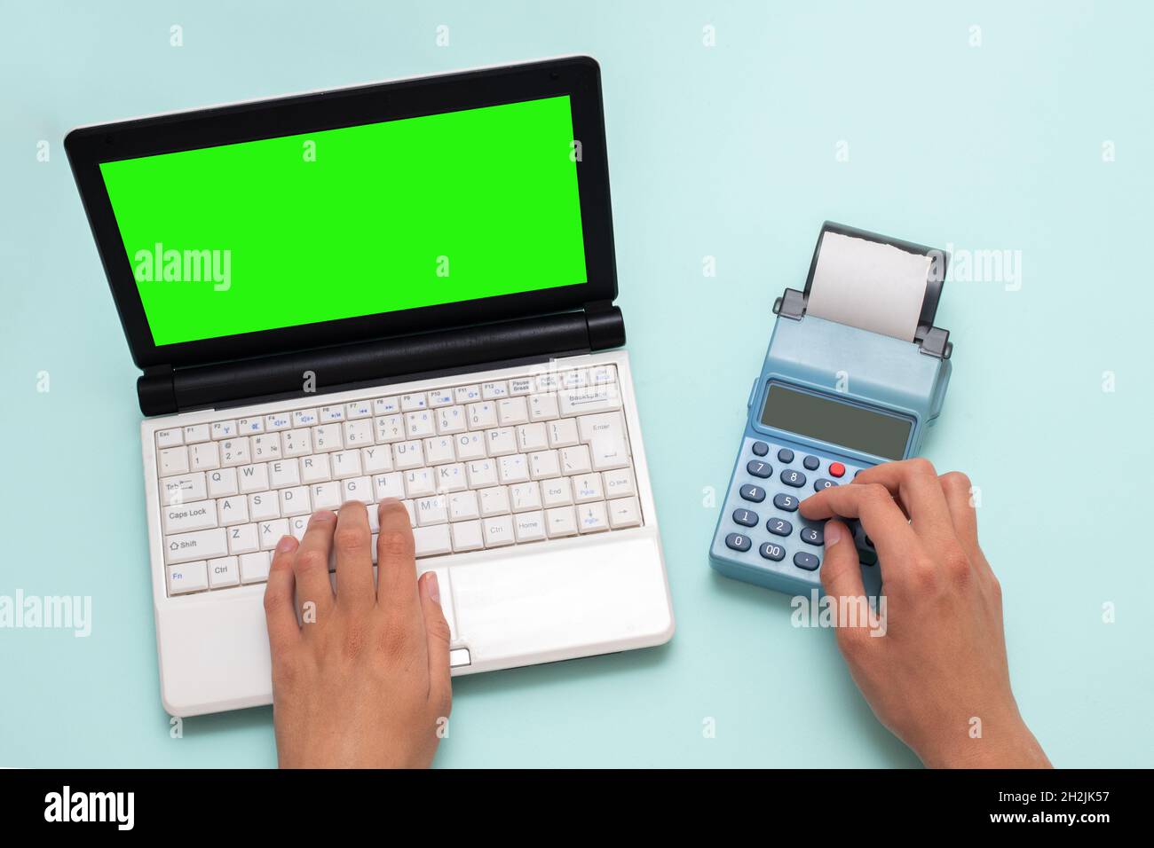 Hand typing on a laptop with a green screen and pressing buttons on a cash register on a blue background, copy space. The cashier selects the item and Stock Photo