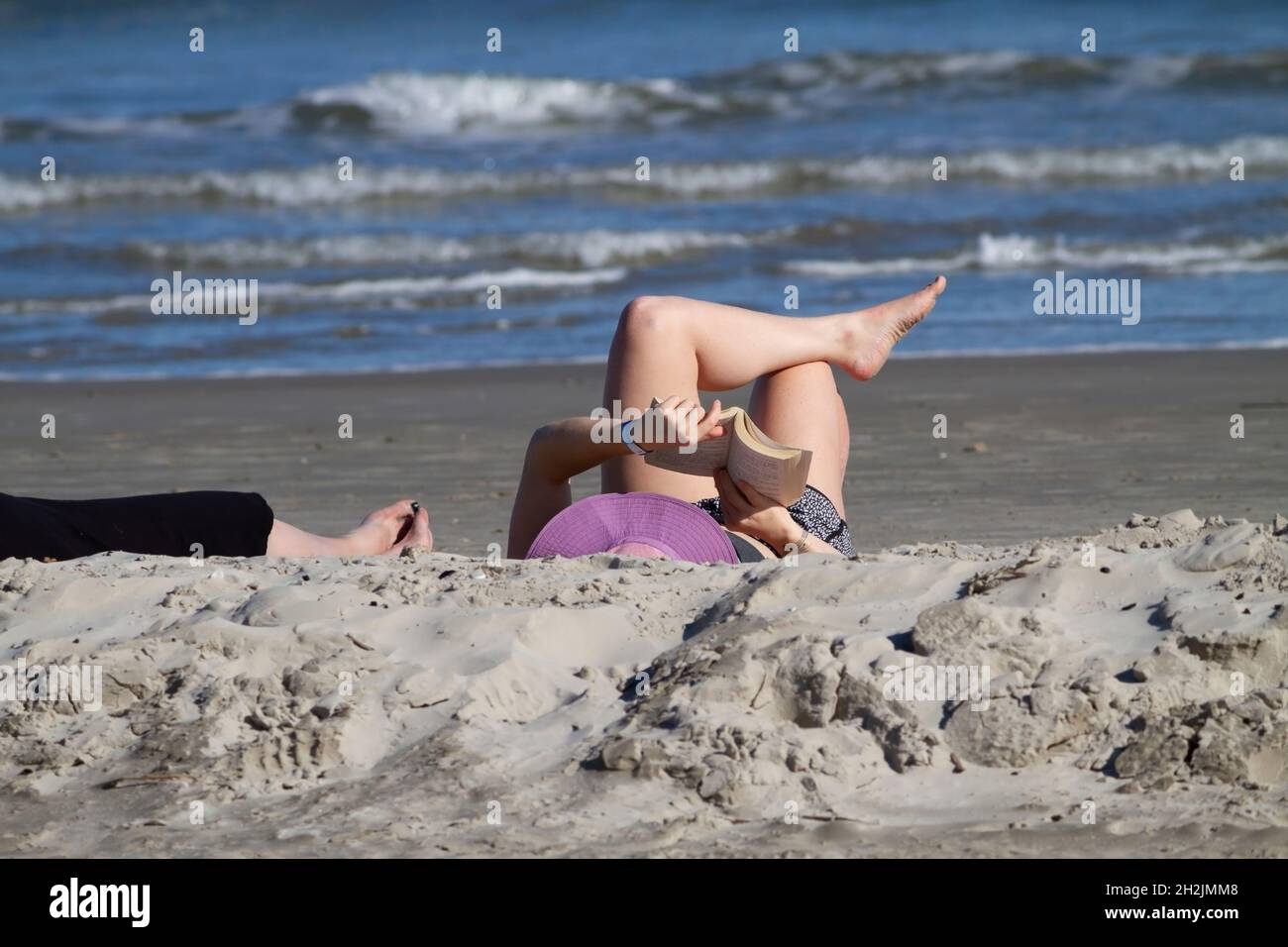Caucasian female reading reclined on Port Aransas, Texas beach. Crossed legs, arms and open book visible. Feet of companion also visible in frame. Stock Photo
