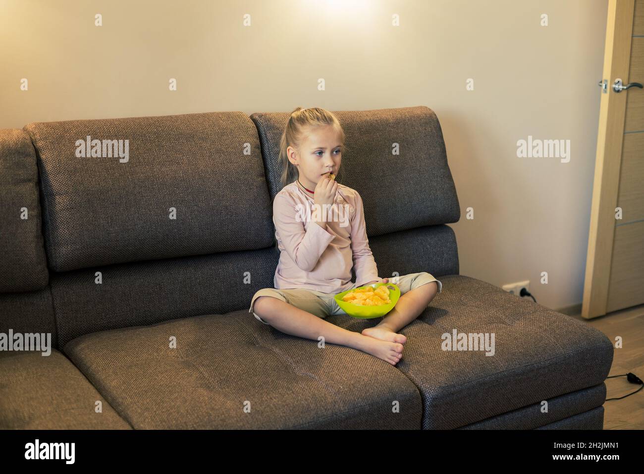 Beautiful little girl eating unhealthy food while watching TV at the sofa Stock Photo