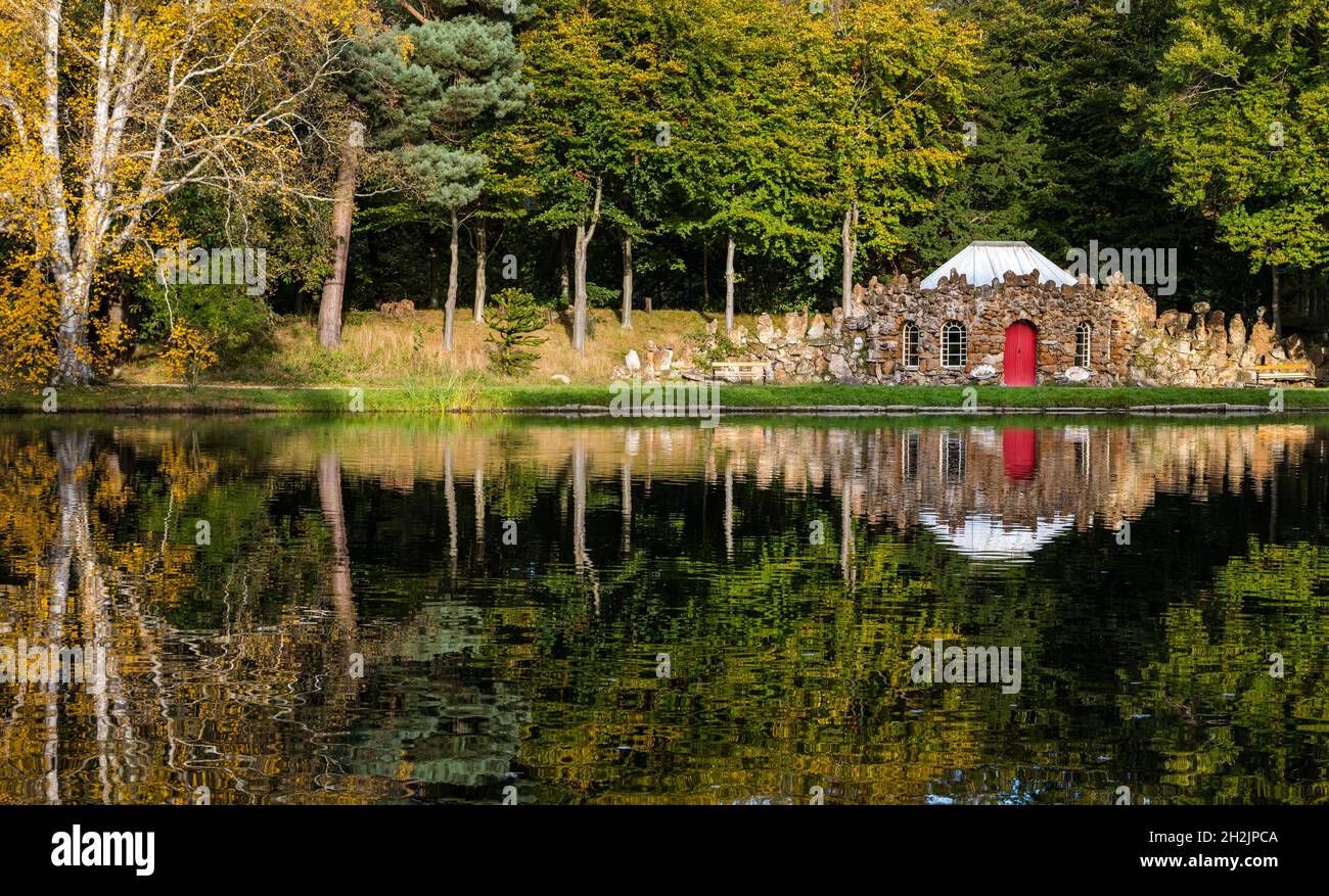 Gosford Estate, East Lothian, Scotland, United Kingdom, 22nd October 2021. UK Weather: Autumn colours: sunshine lights up reflections of the Autumnal trees and the quirky rubble stone curling lodge on the artificial lake in the estate Stock Photo