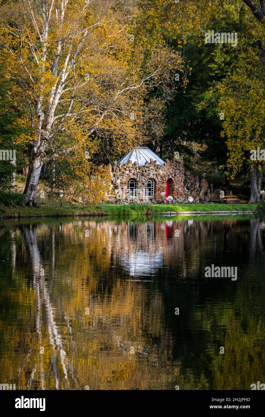 Gosford Estate, East Lothian, Scotland, United Kingdom, 22nd October 2021. UK Weather: Autumn colours: sunshine lights up reflections of the Autumnal trees and the quirky rubble stone curling lodge on the artificial lake in the estate Stock Photo