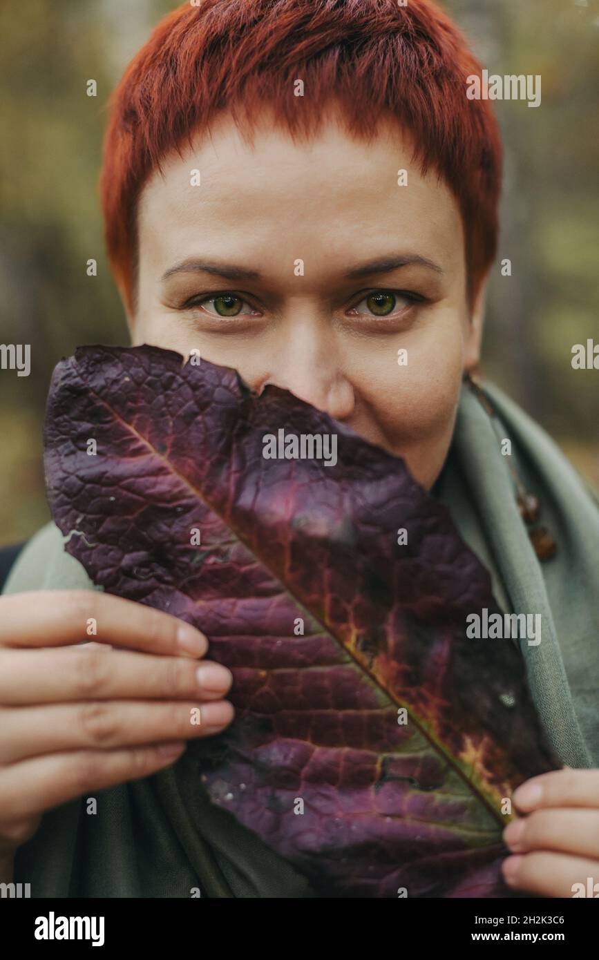 A middle-aged woman of 30-40 years old with an autumn burgundy leaf, her face is covered. Closeup portrait, young adult woman Stock Photo