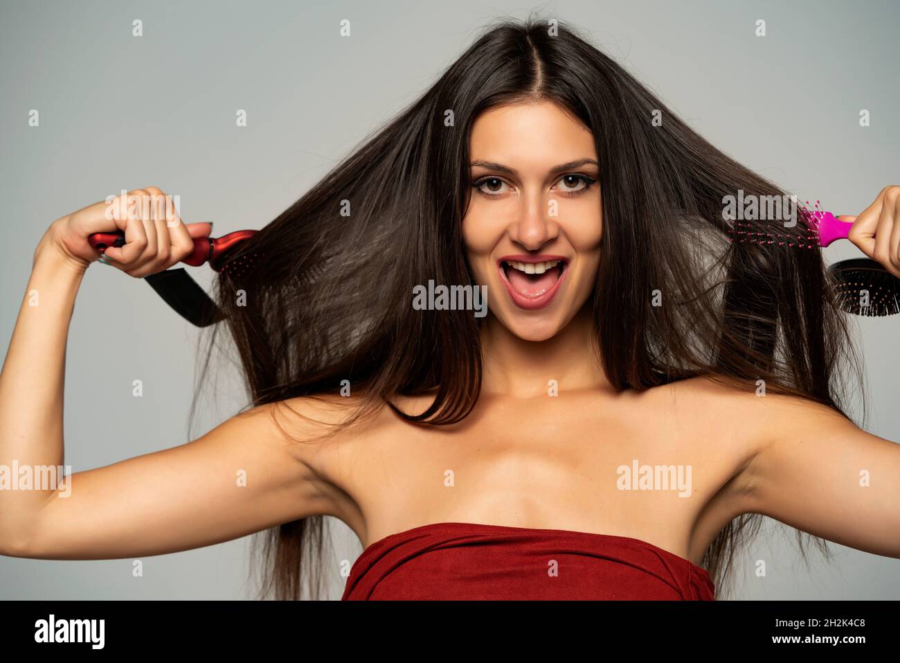 Front view of happy woman brushing her hair with hair brushes on gray