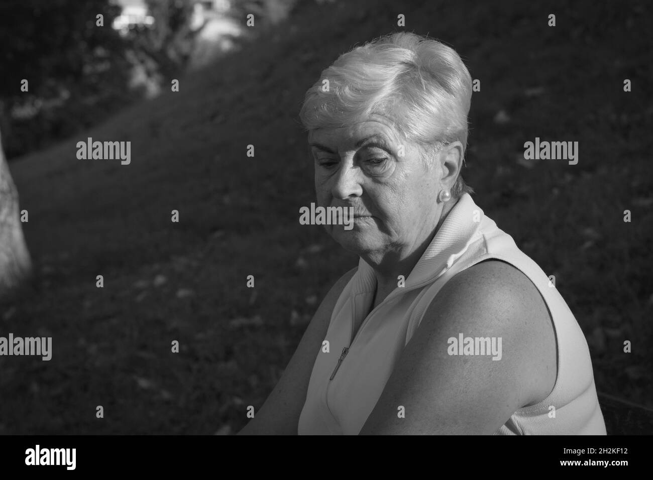Sad senior woman looking down with sunlight on face at park. Depressed elder lady wearing sleeveless white shirt. Pessimistic, worry, serious mother Stock Photo