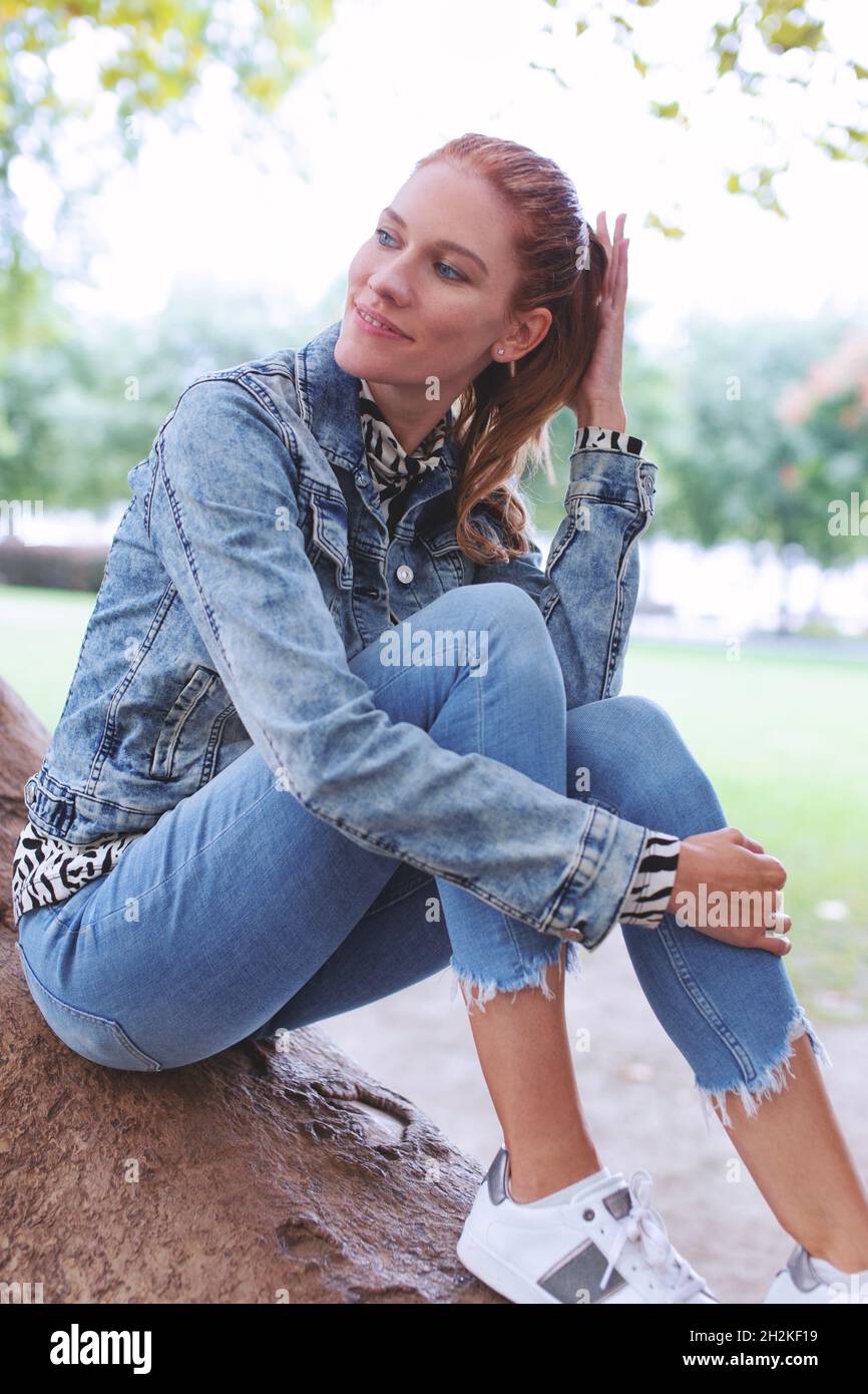 Young natural redhead woman in jeans and denim jacket sitting on branch of tree outdoors Stock Photo