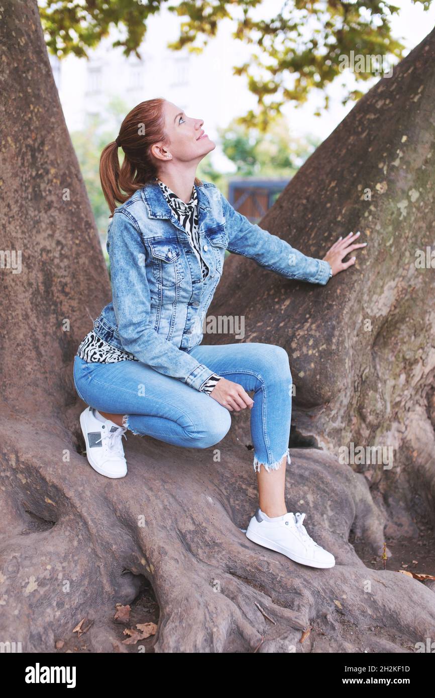 Young redhead Caucasian woman posing on tree and looking up Stock Photo