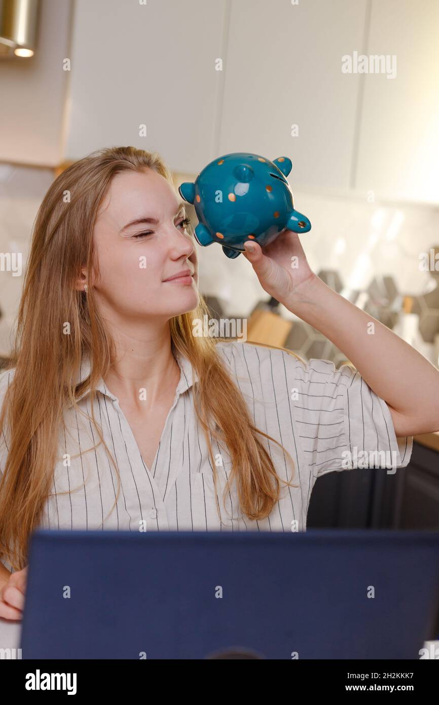 Woman looking at empty piggy bank sitting at home with laptop in kitchen, family budget, personal savings online shopping, credit Stock Photo