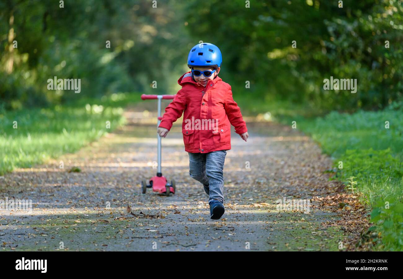 Caucasian child wearing a blue helmet, a red coat and sunglasses, running on a forest path in the fall. A red scooter is in the background; Stock Photo