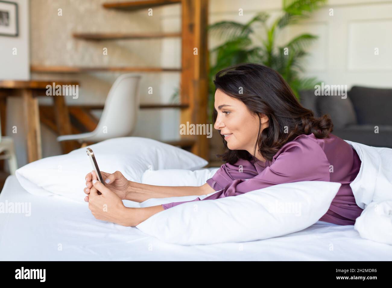 Social media addiction concept - a middle-aged woman checks messages and news on social media right after waking up. She lies in bed and looks at the Stock Photo