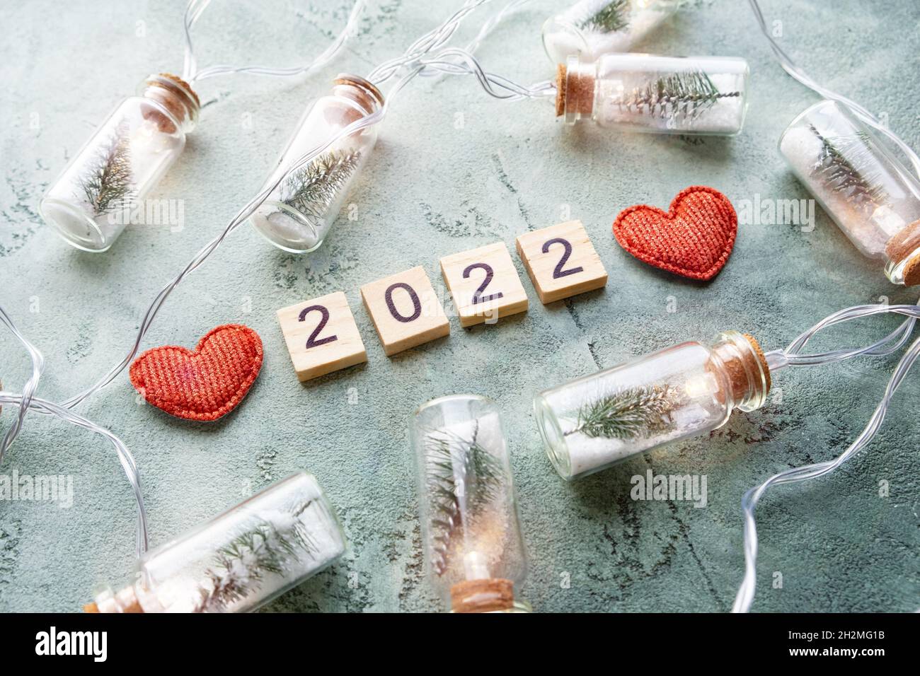 Number 2022 on wooden squares and a glowing garland of glass jars with Christmas trees and snow inside and two hearts on a textured green background Stock Photo