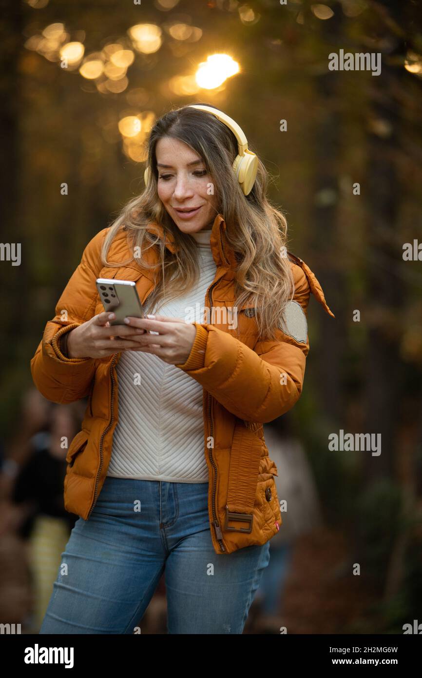 happy blonde woman with headphones and coat typing on her cell phone in the middle of nature Stock Photo