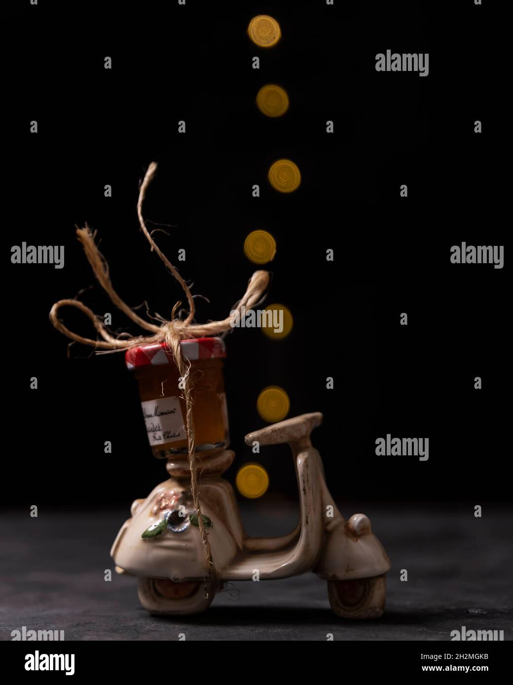 A small glass jar containing peach jam is tied with twine to a model scooter with bokeh lights in the background Stock Photo