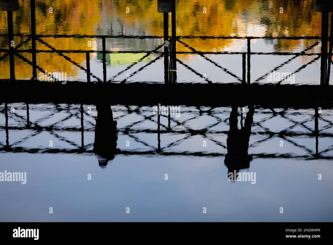 Reflection in water of people walking over a pedestrian bridge in a park during a sunny autumn day. Stock Photo