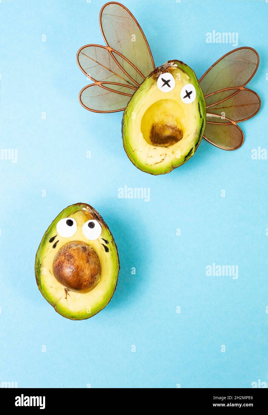 2 halves of avocado fruit making faces (dead and scared), with dragonfly wings on bright blue background. Stock Photo