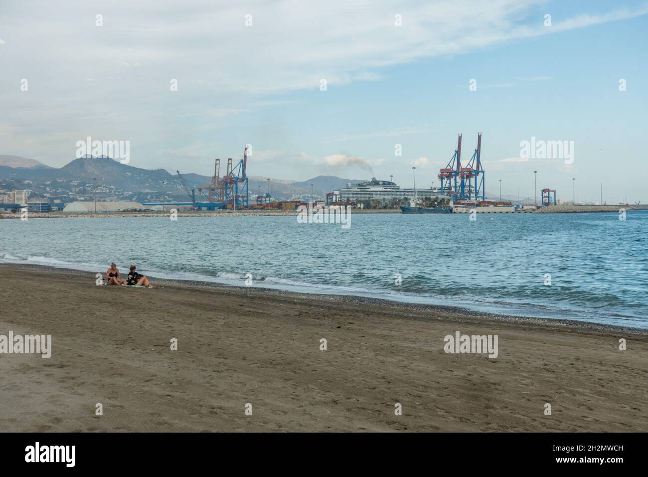 People on Huelin beach in autumn, with the industrial setting cargo port and cranes behind, Malaga, Costa del sol, Spain. Stock Photo