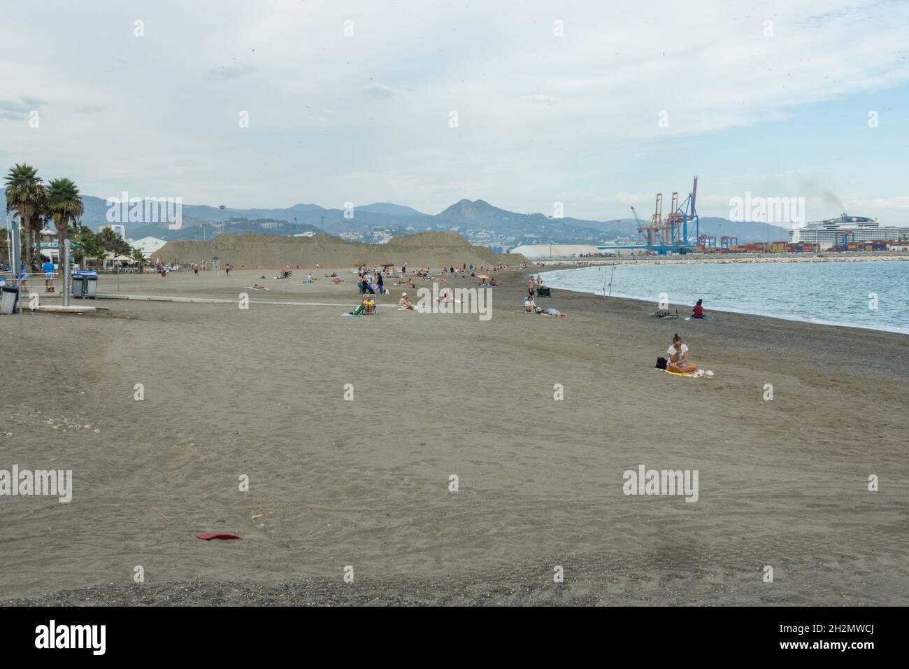 People on Huelin beach in autumn, with the industrial setting cargo port and cranes behind, Malaga, Costa del sol, Spain. Stock Photo