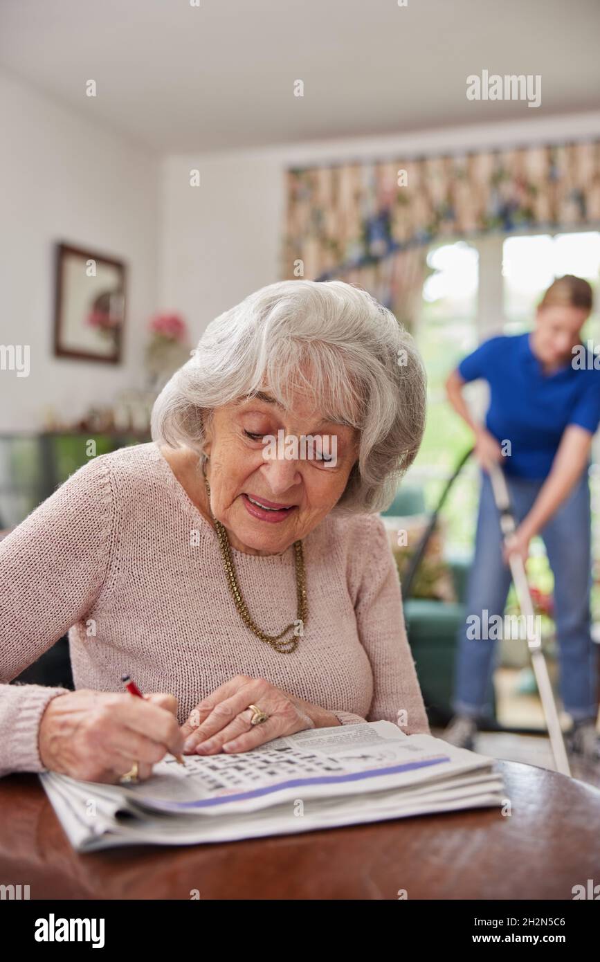 Female Home Help Cleaning House With Vacuum Cleaner Whilst Senior Woman Does Crossword In Newspaper Stock Photo