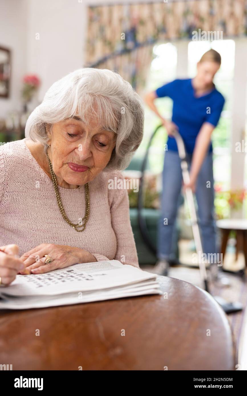 Female Home Help Cleaning House With Vacuum Cleaner Whilst Senior Woman Does Crossword In Newspaper Stock Photo