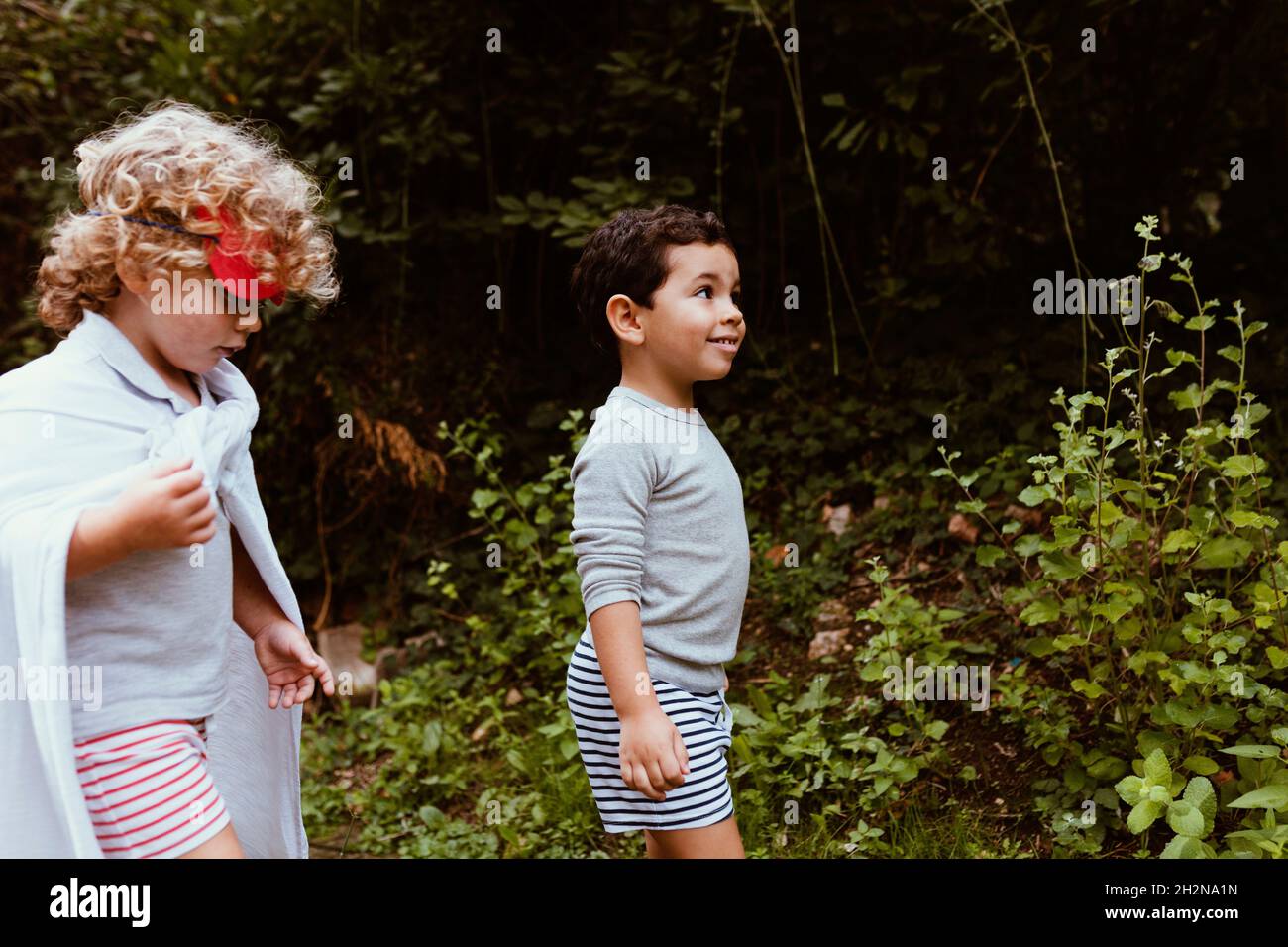 Blond boy wearing cape walking with male friend in forest Stock Photo