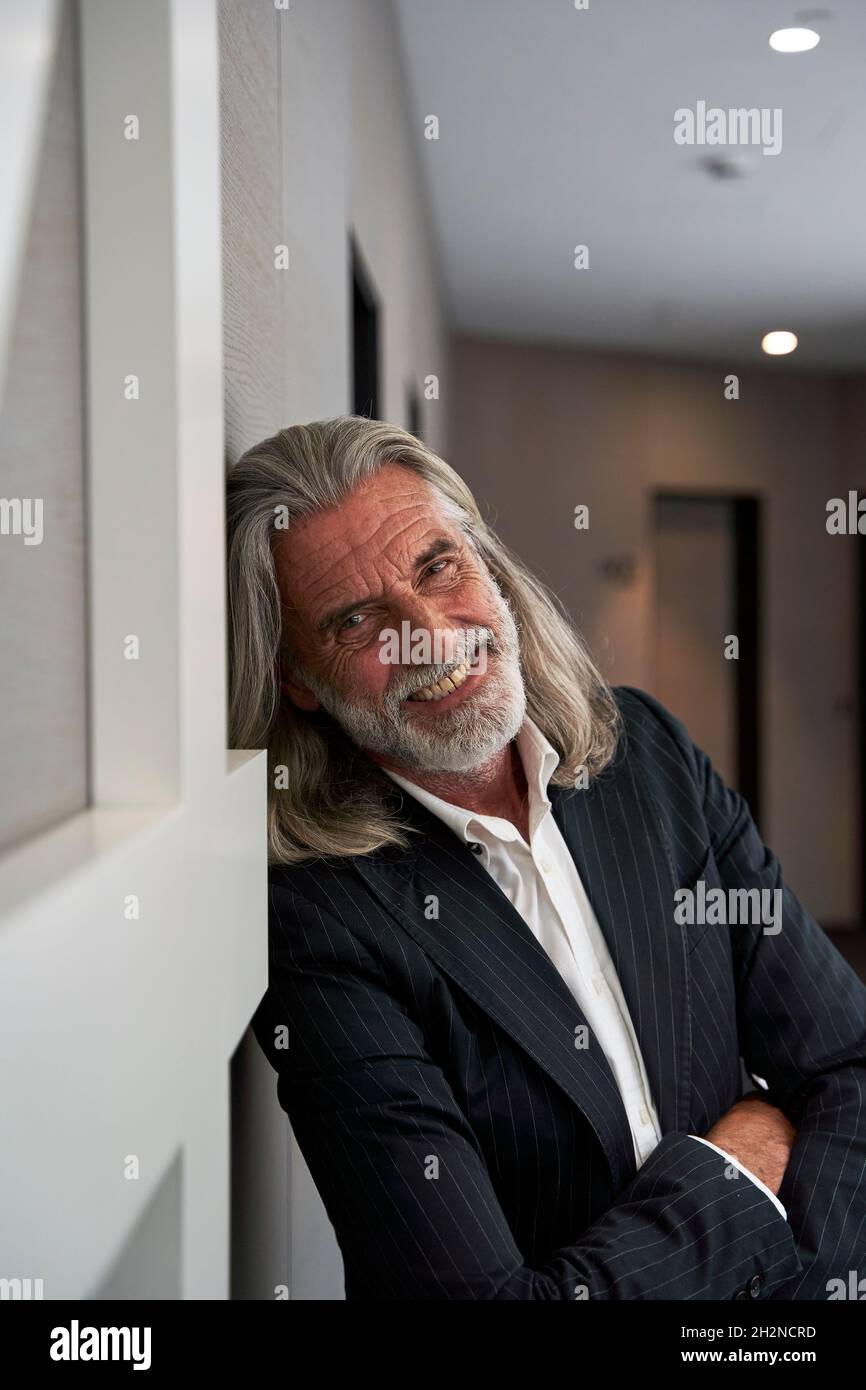 Smiling businessman with arms crossed leaning on wall at hotel corridor ...