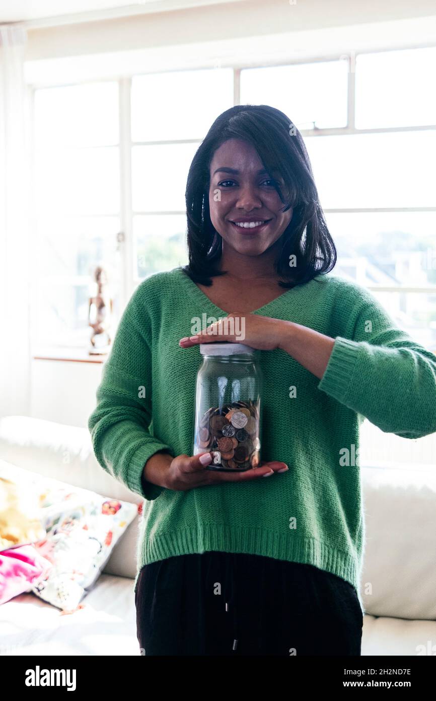 Smiling woman holding coin's jar in living room Stock Photo