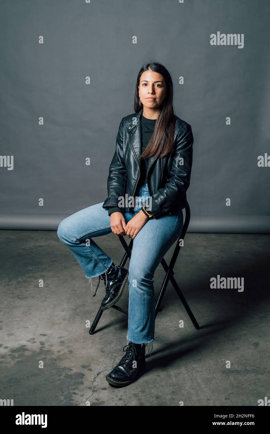 Young confident woman in leather jacket sitting on chair in studio ...