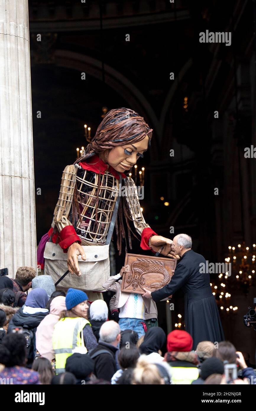 London, UK. 23rd Oct, 2021. The Pastor from the St Paul Cathedral receives the token of appreciation from the Little Amal.Little Amal, a 3.5 meters tall puppet portrayed as a 9 year old Syrian refugee girl and representing all refugee children, arrived central London at the Great Door of the St Paul Cathedral. She has been on journey since July this year departing from the Syrian-Turkish border in search of her 'mother'. She will walk 8000 km and arrive at Manchester in early November as her final destination, where it is also the location of the short term holding facility for refugees in the Stock Photo