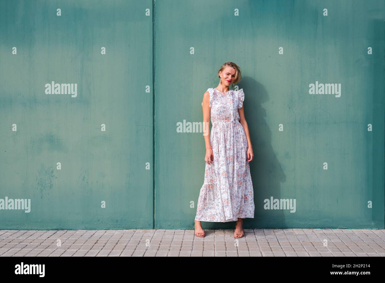 horizontal portrait of a blonde thin woman in floral summer dress. She has is leaning on a green wall. Stock Photo
