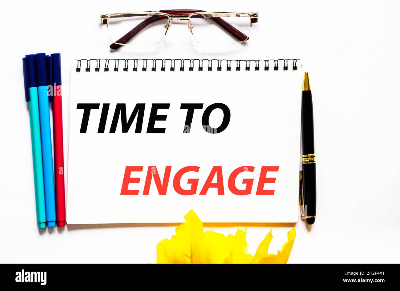 Time to engage, the text is written on a notebook, next to glasses, a pen and felt-tip pens on a white background. Stock Photo
