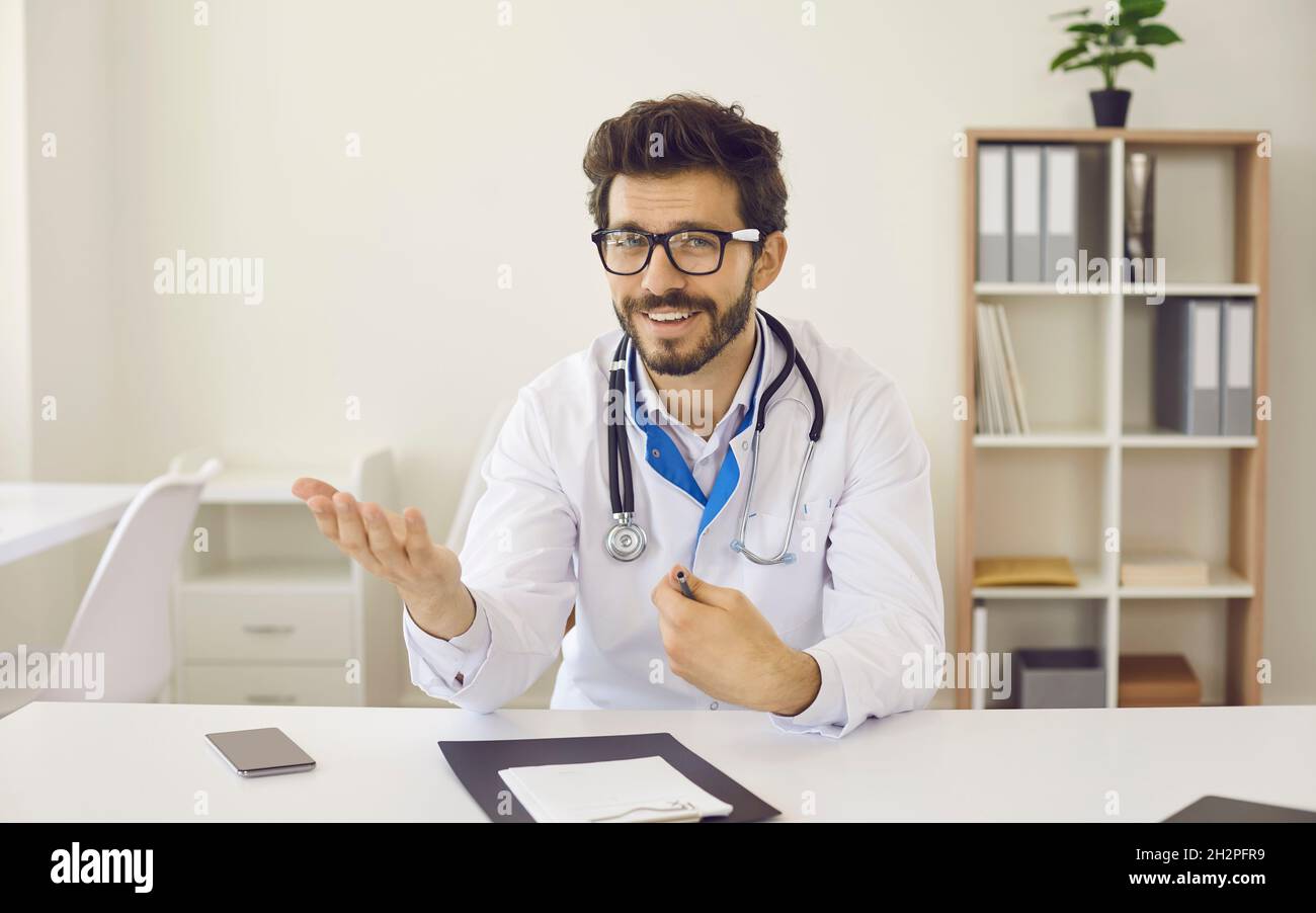 Young friendly male doctor sits in front of webcam and conducts online consultation with patient. Stock Photo