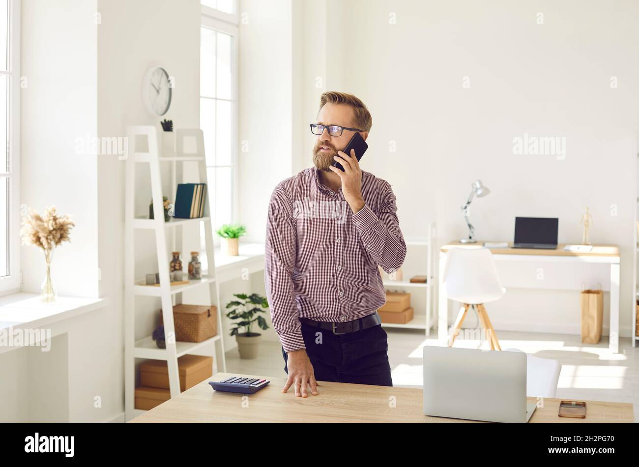 Serious businessman standing by office desk with laptop and making call ...