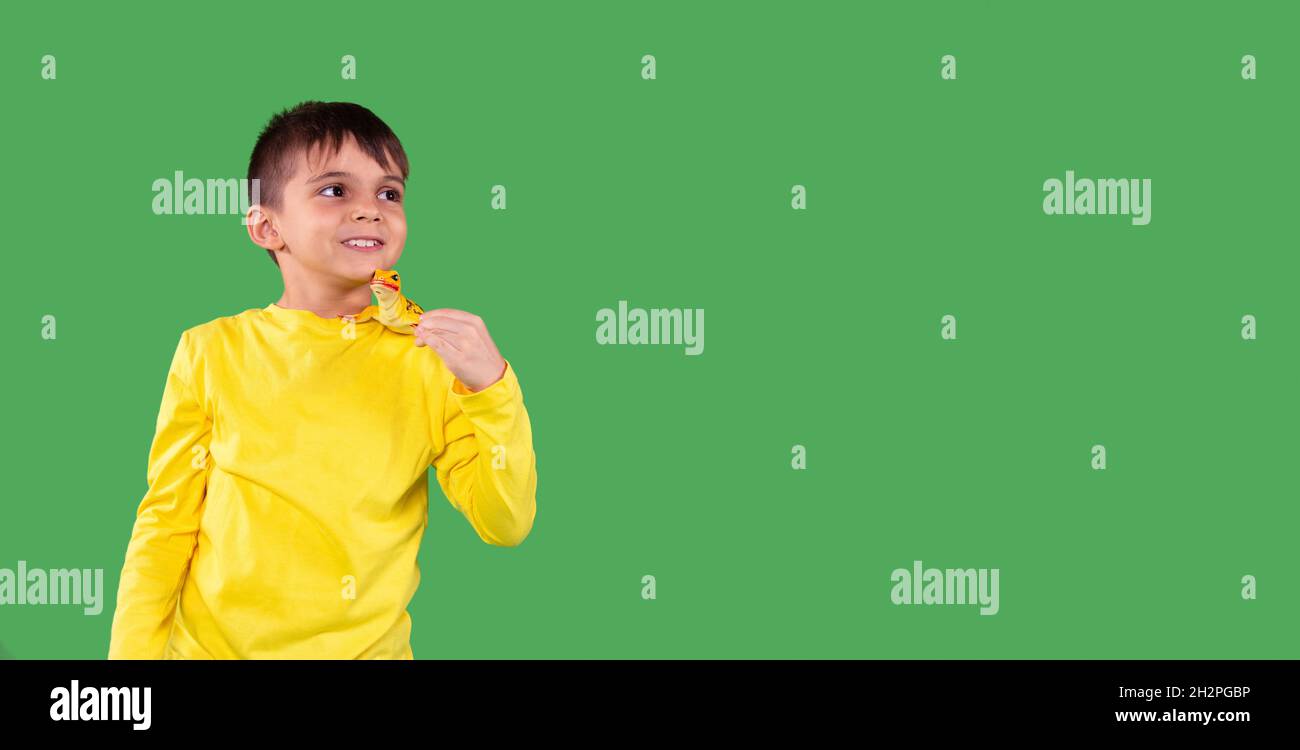 Photo of a smiling school aged boy in yellow hoodie looking aside and holding the dinosaur toy on his shoulder against the green background, empty Stock Photo