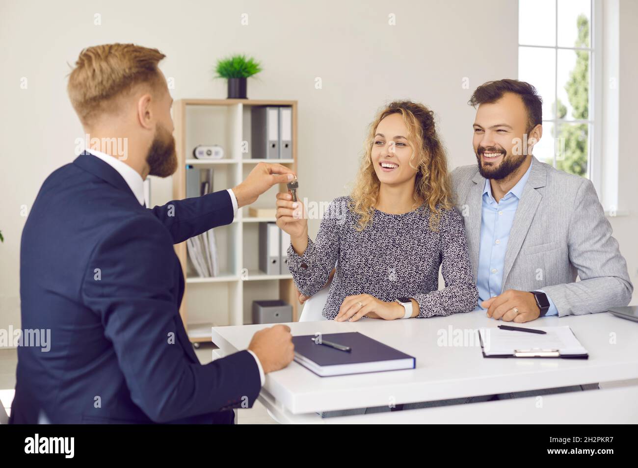 Real estate agent hands over keys to his new home to excited married couple at meeting in office. Stock Photo