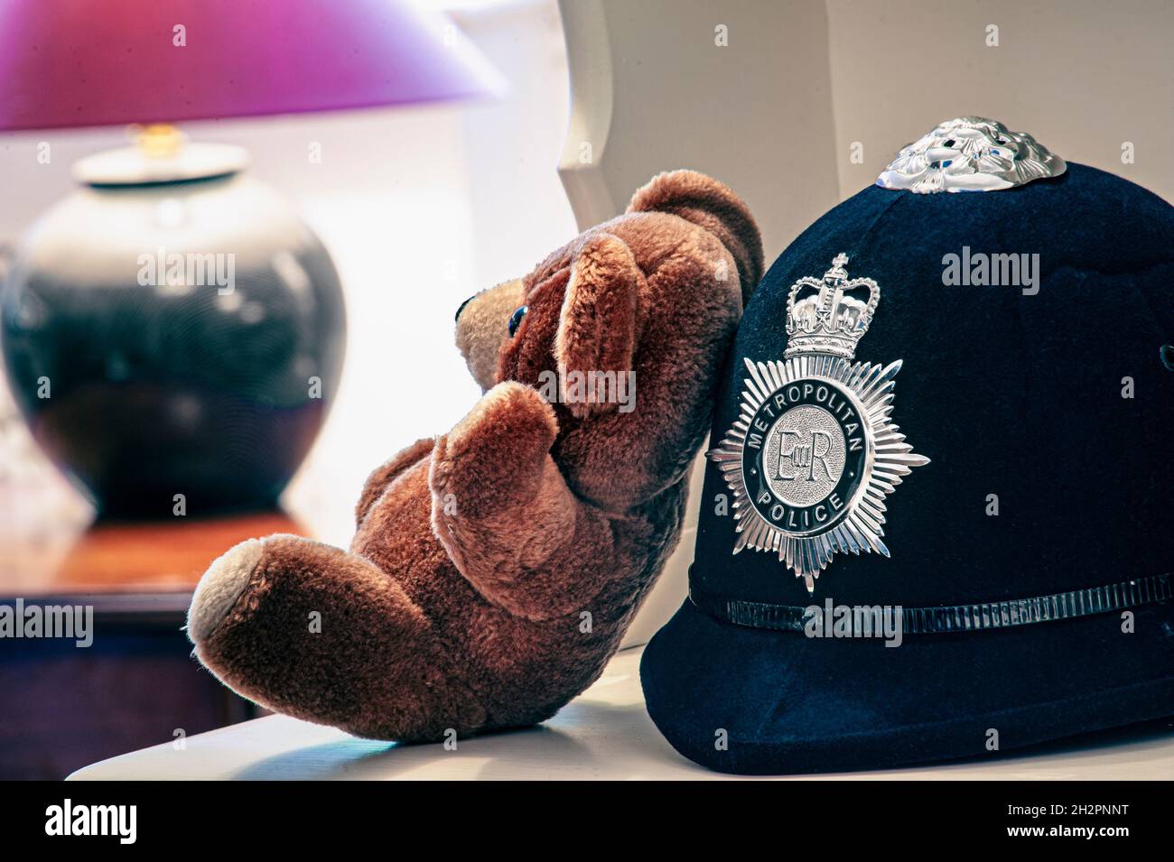 Policeman Police Trust Helmet Home Children Child Infant Concept faith honesty trust trusting image of Metropolitan Policeman's helmet with child's teddy bear leaning against it in a domestic home situation. Stock Photo