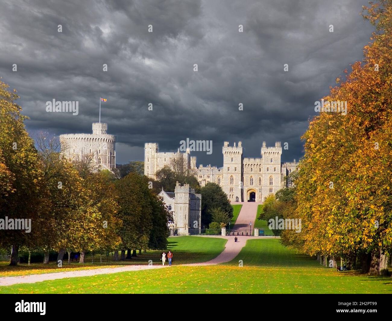 Windsor Castle with stormy sky flying the Royal Standard viewed down the Long Walk illuminated by dramatic sunlight with dark brooding storm clouds and autumnal colour Berkshire UK Stock Photo