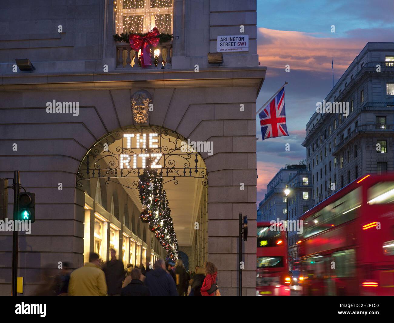 RITZ LONDON CHRISTMAS CROWDS The Ritz Hotel at winter busy festive season, evening night lights ‘The Ritz’ sign illuminated, with a Union Jack flag, shoppers and passing blurred London red buses Arlington Street Piccadilly London UK Stock Photo