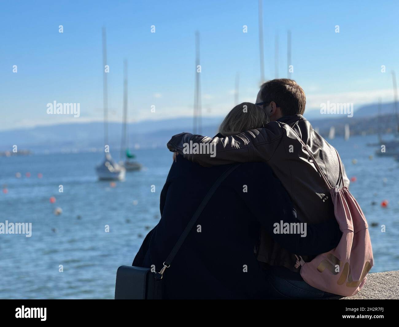 Young heterosexual couple cuddling together in back view. They are sitting on a concrete barrier observing lake Zurich with boats on it. Stock Photo