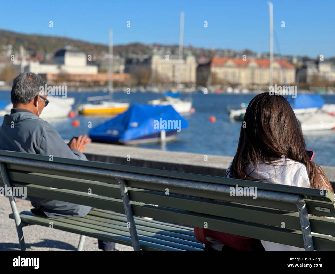 Woman and man sitting on a wooden bench and keep social distancing.  The sit on opposite ends of bench and each of them is looking into their phones. Stock Photo