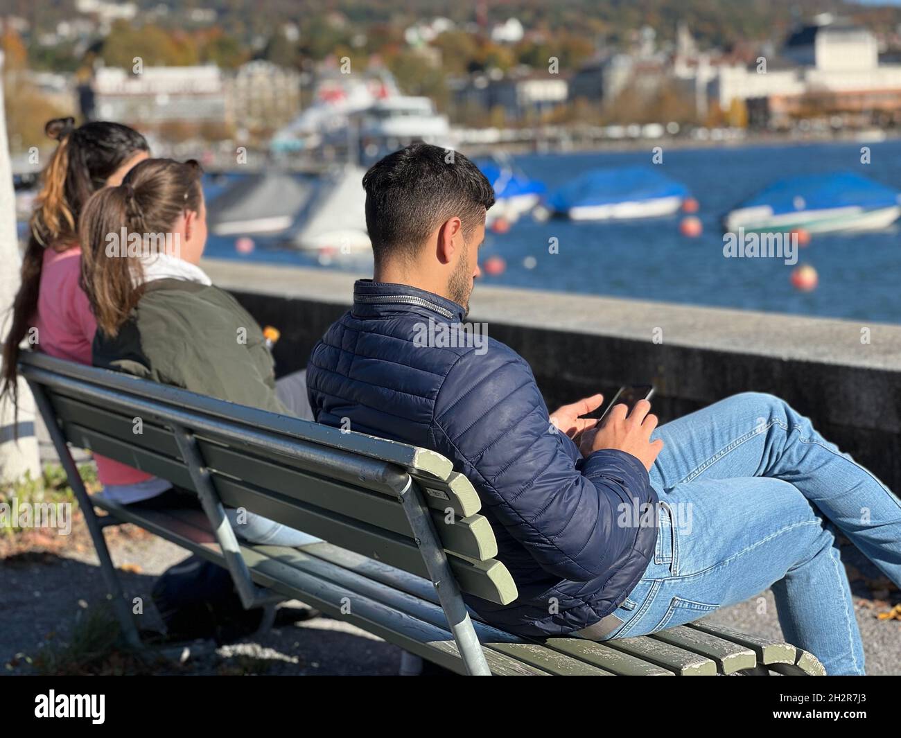 Two women and one man sit on wooden bench and keep social distancing.  They sit on opposite ends of bench and each of them look into their cell phones Stock Photo
