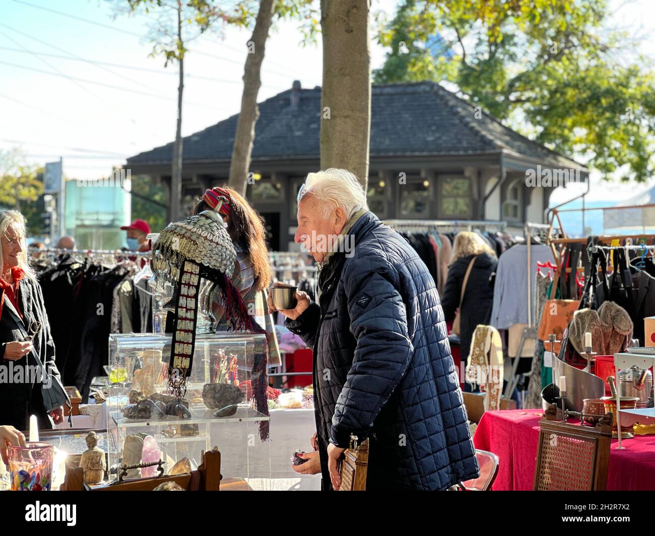 Vendor at flea market is drinking coffee from a flask. He is at his stall selling vintage and rare merchandise. Elderly man is captured in side view. Stock Photo