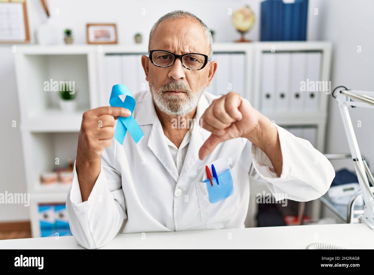 Mature doctor man holding blue ribbon at the clinic with angry face ...