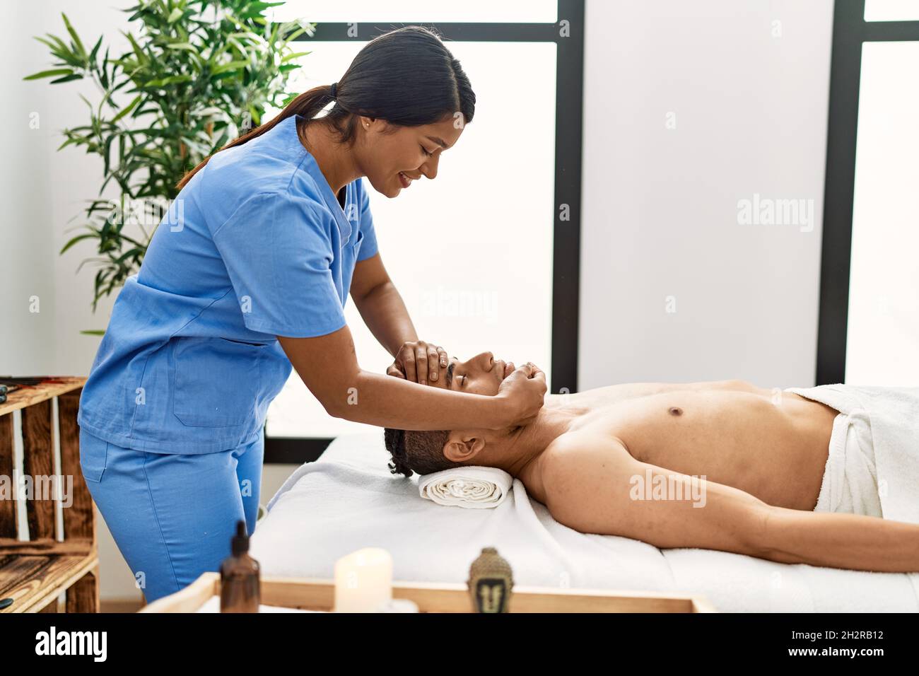 Young latin massage therapist woman giving relaxing treatment on man head at beauty center. Stock Photo