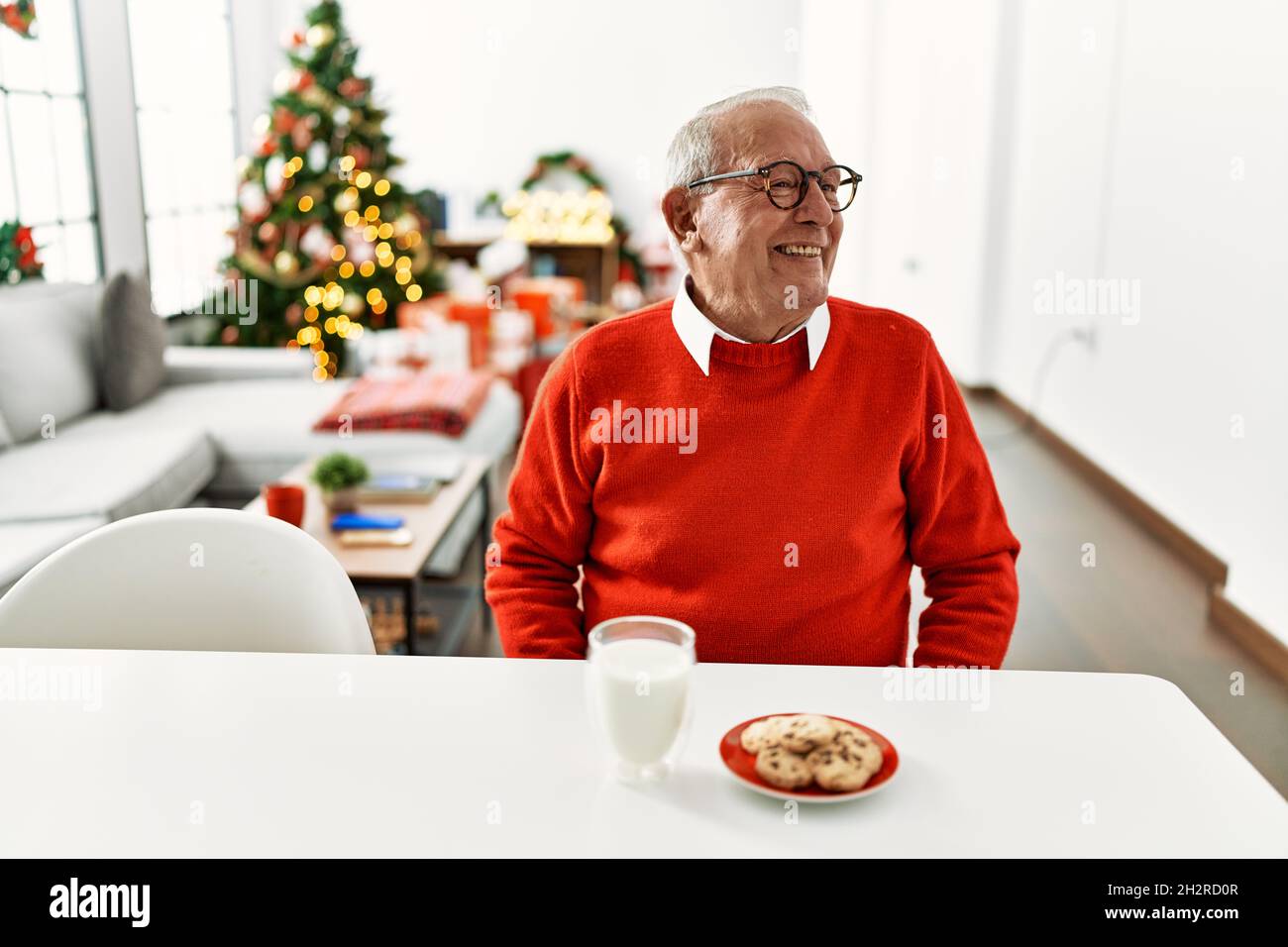 Senior man with grey hair sitting on the table with cookies by christmas tree looking away to side with smile on face, natural expression. laughing co Stock Photo