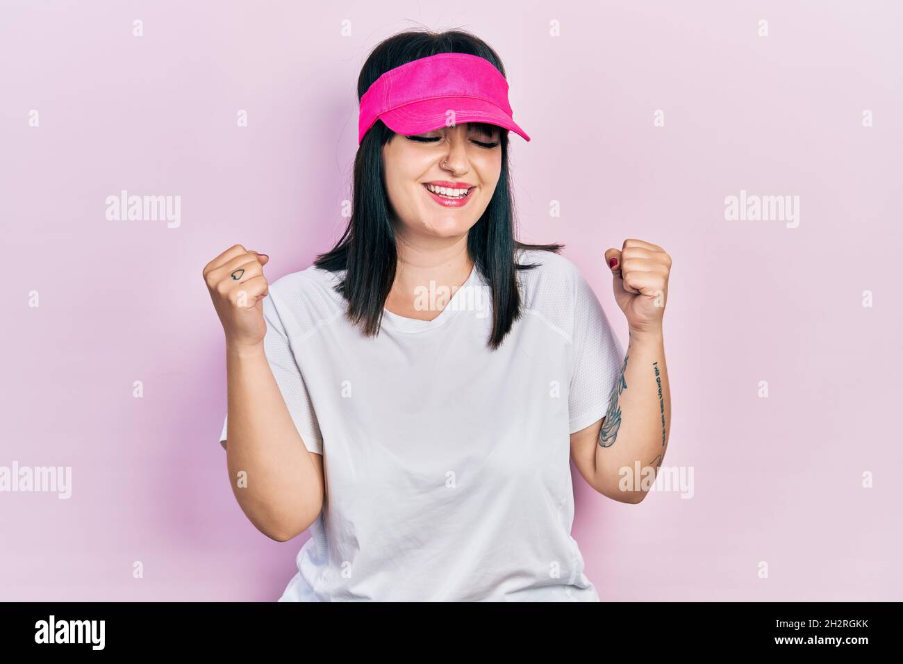 Young hispanic woman wearing sportswear and sun visor cap very happy and excited doing winner gesture with arms raised, smiling and screaming for succ Stock Photo
