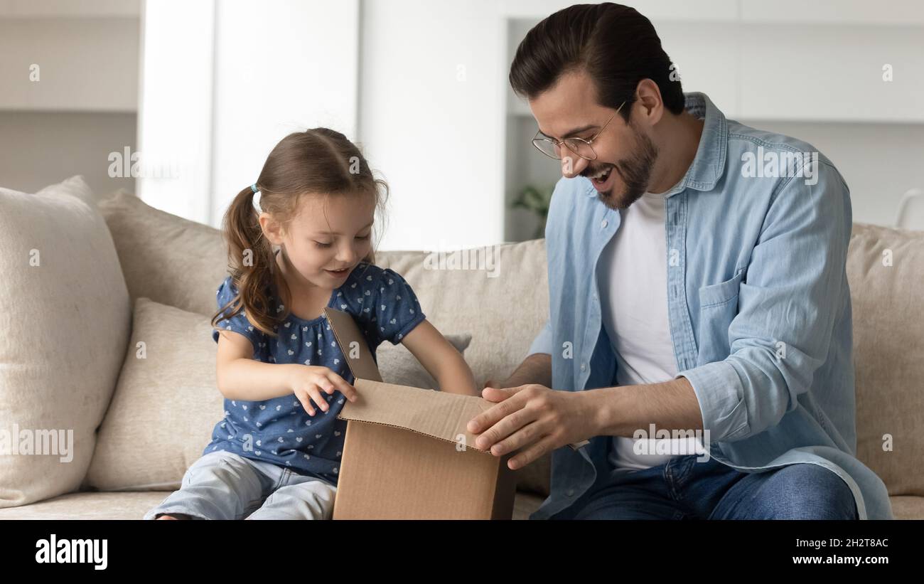 Joyful adorable small child girl opening carton parcel with happy father. Stock Photo