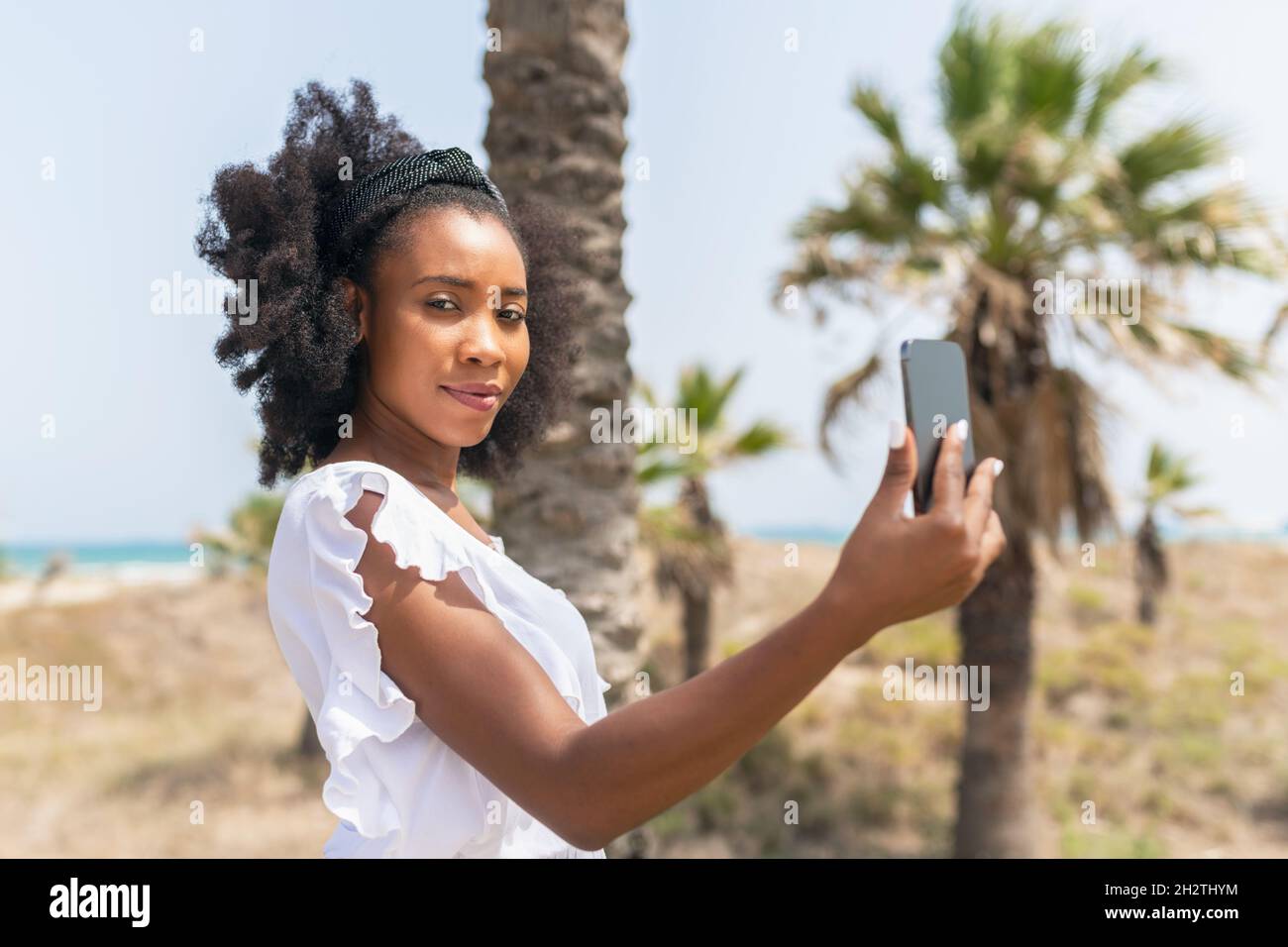 African woman facing the camera while holding a smartphone in a beach in a windy day Stock Photo