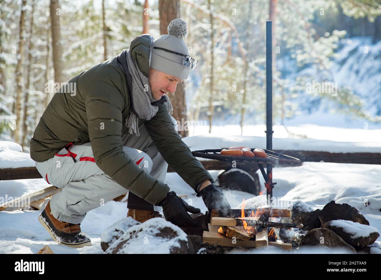 Man roasting sausages on campfire in forest by the lake, making a fire, grilling. Happy tourist exploring Finland. Beautiful sunny winter landscape Stock Photo